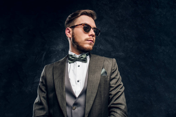 A stylish young man in sunglasses dressed in an elegant suit against a dark textured wall - Photo, Image