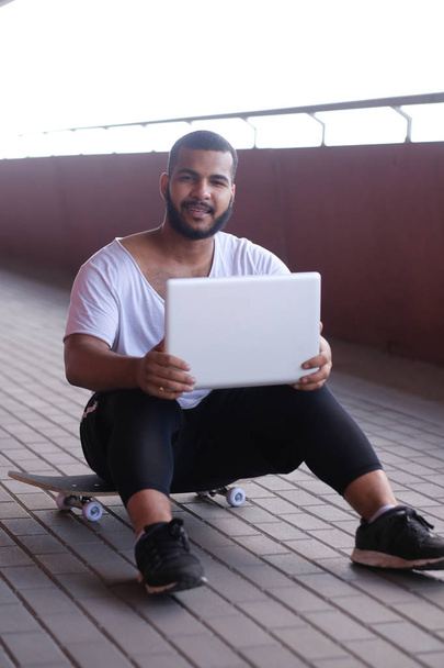 El afroamericano está sentado en la acera de un puente, sosteniendo el portátil de rodillas y mirando una cámara
 - Foto, imagen