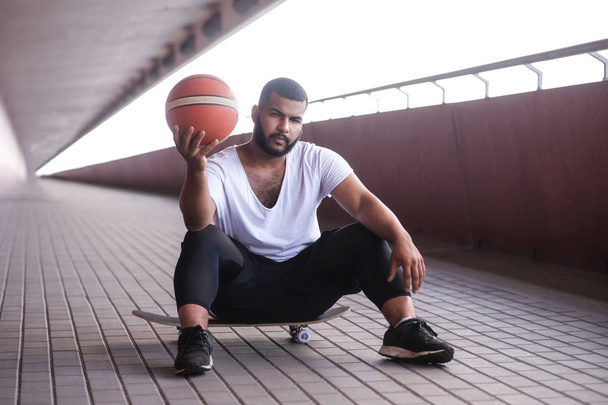 Hombre afroamericano está sentado en un monopatín y sosteniendo una pelota de baloncesto en una acera de puente
 - Foto, imagen