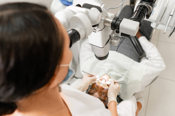 Focus on microscope. Young woman dentist treating root canals using microscope in the dental clinic. Man patient lying on dentist chair with open mouth. - Photo, image