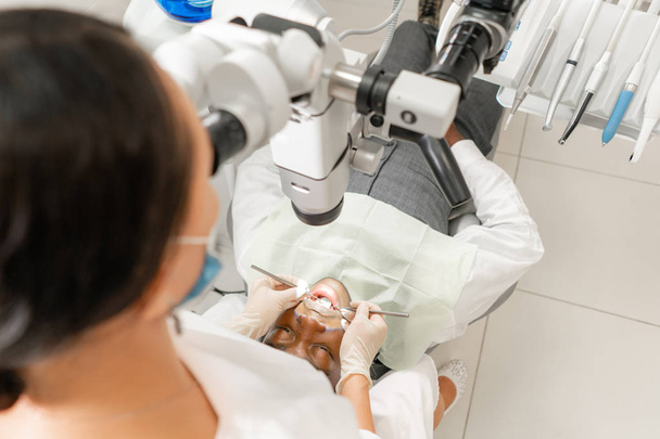 患者に焦点を当てます。若い女性の歯科医が歯科医院での顕微鏡を用いた根管治療します。口を開けて歯科医の椅子に横になっている男性患者. - 写真・画像