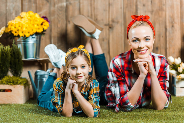 cheerful mother and daughter lying with crossed legs on grass near wooden fence and flowers - Photo, Image