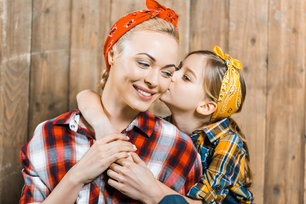cute daughter kissing cheek of happy mother near wooden fence  - Foto, imagen