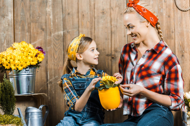 Tochter blickt fröhliche Mutter an, während sie Blumen am Holzzaun berührt  - Foto, Bild
