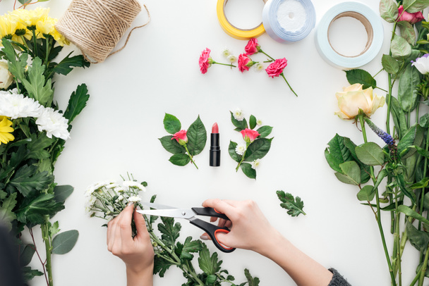 bijgesneden beeld van vrouwelijke overhandigt snijden chrysanten met boutonnieres en lippenstift omringd door bloemen op witte achtergrond - Foto, afbeelding