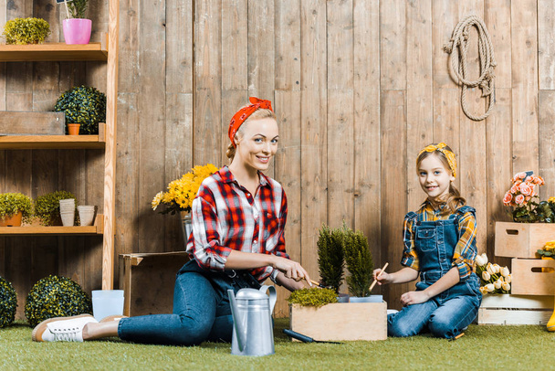 mother planting flowers with cute daughter while sitting on grass near wooden fence - Photo, Image