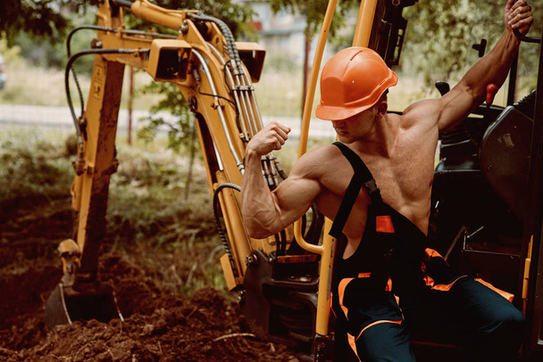 Employee concept. Employee flex biceps and triceps muscle. Strong employee at excavator cabin. Employee in working uniform on excavation site. Strength and masculinity - Φωτογραφία, εικόνα