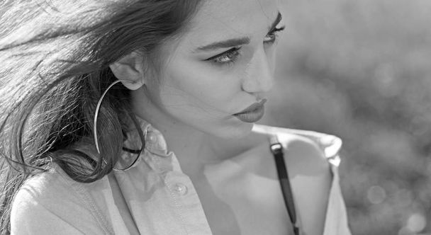Woman on calm face with make up, nature on background, defocused. Girl with big rings earrings wears unbuttoned shirt and black lingerie. Lady with nude shoulder looks attractive. Femininity concept. - Photo, Image
