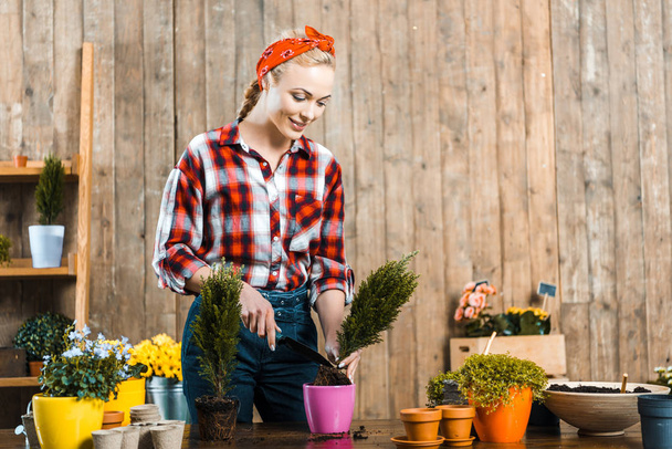 fürsorgliche Frau lächelt, während sie Schaufel hält und Pflanze in Topf pflanzt  - Foto, Bild