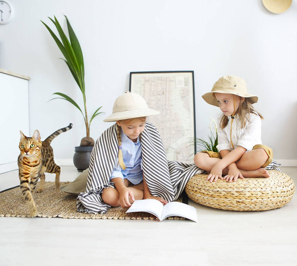 Two little girls are playing at home to travelers with bengal cat wearing vintage style clothes. Childhood, fantasy, imagination - Photo, Image