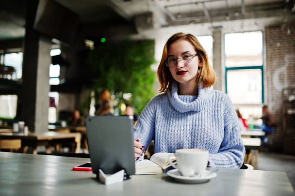 Cheerful young beautiful redhaired woman in glasses using her phone, touchpad and notebook while sitting at her working place on cafe with cup of coffee. - Photo, image