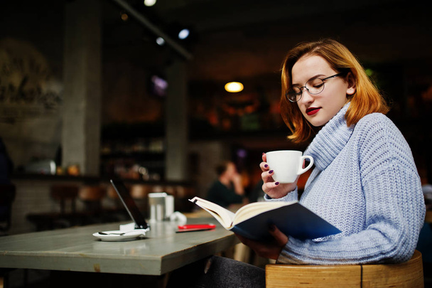 Cheerful young beautiful redhaired woman in glasses using her phone, touchpad and notebook while sitting at her working place on cafe with cup of coffee. - Φωτογραφία, εικόνα