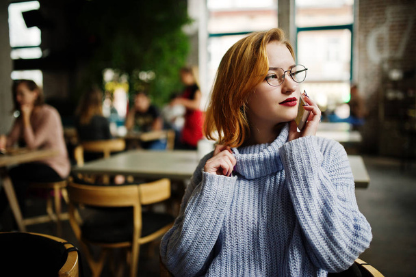 Vrolijke jonge mooie roodharige vrouw in glazen zittend op haar werkplek op café en spreken door cellphone. - Foto, afbeelding