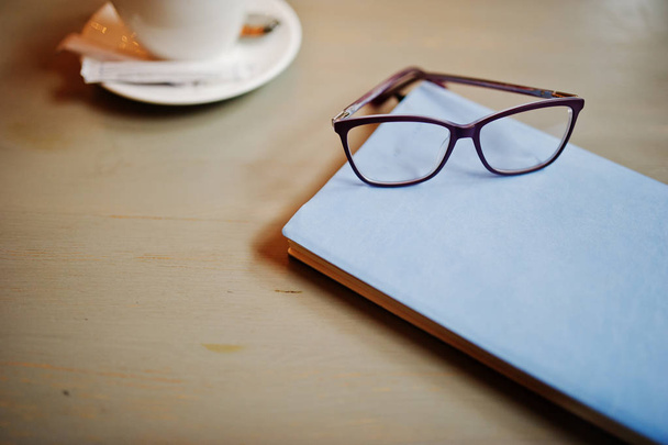Close up photo of notebook with glasses against cup of coffe at cafe table. Eyewear concept. - Fotografie, Obrázek