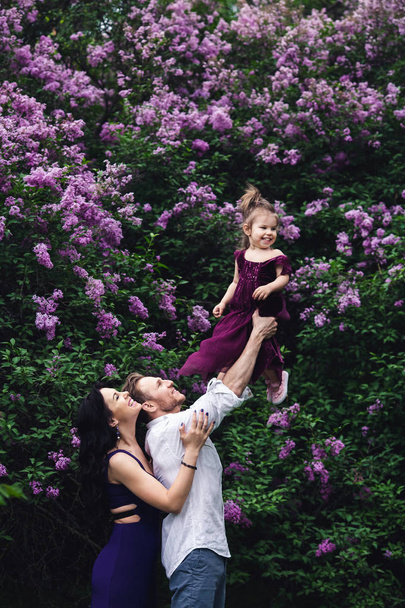 vrolijke en gelukkige familie in de buurt van lila bloeiende struiken. vader gooit van dochtertje omhoog. - Foto, afbeelding