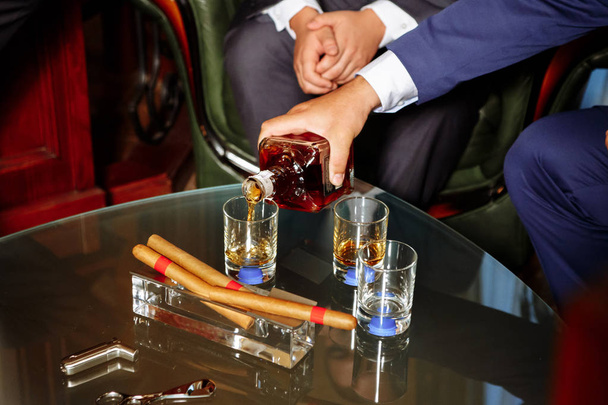 mies kaataa alkoholia kolmeen lasiin, kolme sikaria makaa tuhkakupissa
 - Valokuva, kuva