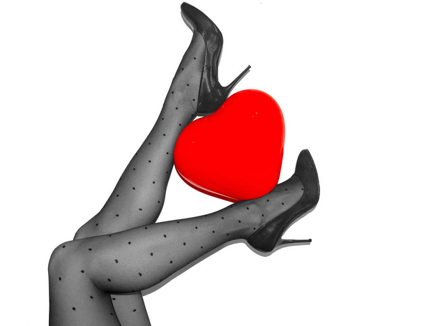 γυναικεία πόδια σε μαύρο οπάκ καλσόν και βυσσινί υψηλής φτέρνα παπούτσια κρατώντας ballon κόκκινη καρδιά σε άσπρο φόντο - Φωτογραφία, εικόνα
