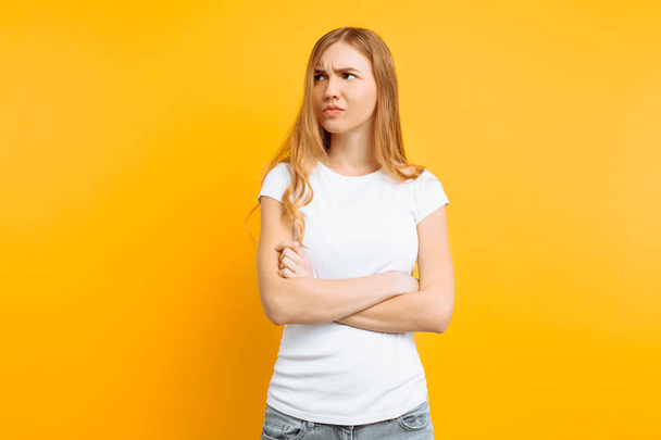 Portrait of an upset young girl, in a white T-shirt, standing with folded hands and offended face, on a yellow background - Foto, afbeelding