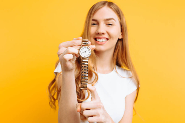 Portrait of a positive young woman, in white t-shirt, pointing her finger at the clock, on a yellow background - Foto, Imagem