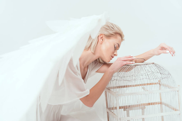 красивая женщина в костюме ангела с крыльями, опираясь на птичью клетку, изолированную на белом
 - Фото, изображение