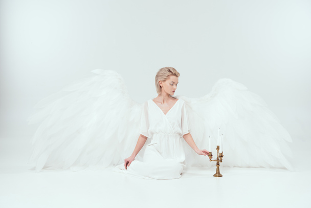 piękna kobieta w strój anioła ze skrzydłami, siedzi w pobliżu Kandelabr ze świecami na białym tle - Zdjęcie, obraz