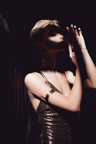 beautiful sexy woman with angel wings and golden accessories posing and gesturing with hands isolated on black - Photo, Image