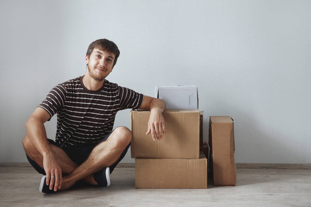 Jeune homme assis sur le sol dans un nouvel appartement après réparation parmi les boîtes en carton
 - Photo, image