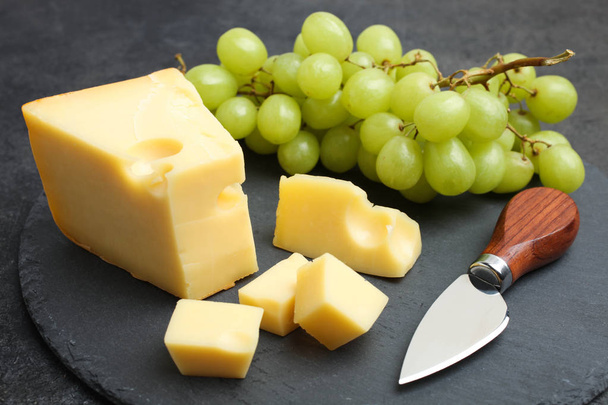 Queso de Maasdam amarillo en rodajas duras con uvas blancas y cuchillo de queso en pizarra sobre fondo negro. Concepto de servir queso
. - Foto, imagen
