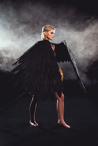 beautiful woman with black angel wings and sword posing on black background - Fotó, kép