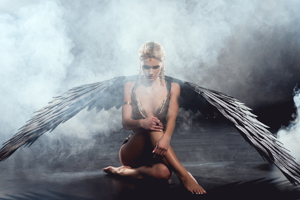 hermosa mujer sexy con alas de ángel negro sentado y posando sobre fondo oscuro
 - Foto, imagen