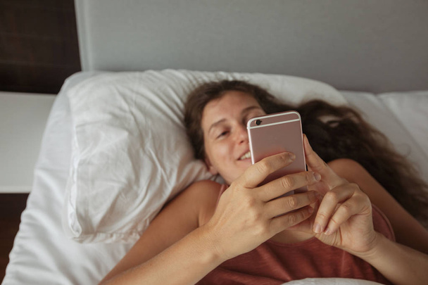 Girl with long hair lies bed looks into phone - Foto, imagen