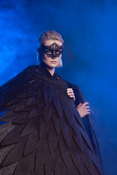beautiful mysterious woman with lace mask and black angel wings looking at camera on dark blue background - Photo, Image