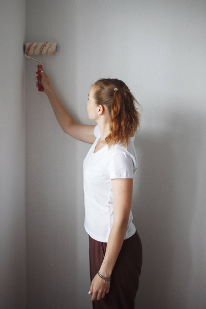 Νεαρή γυναίκα στο λευκό T-shirt ζωγραφική τοίχου με ρολό σε νέο διαμέρισμα - Φωτογραφία, εικόνα