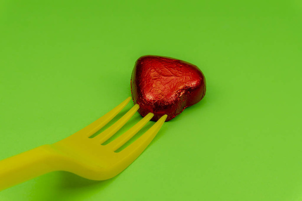 Caramella al cioccolato a forma di cuore in lamina rossa e forchetta di plastica monouso gialla, vista in primo piano su sfondo verde. Il concetto di spezzare i cuori da donnaiolo o seduttore e tradimento
 - Foto, immagini