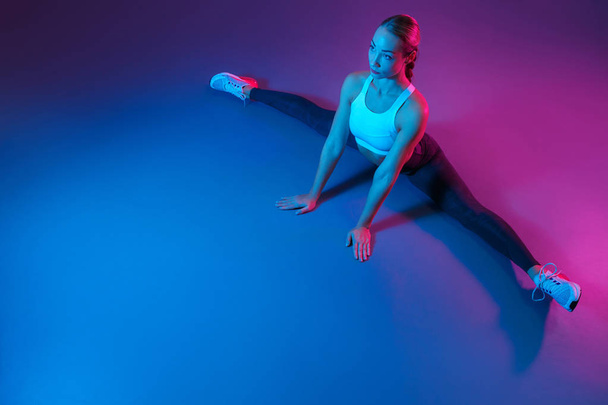 Attractive young slim gymnast woman in sports clothing stretching on the floor in neon lights. Flexible muscular woman doing gymnastic and yoga split. - Foto, immagini