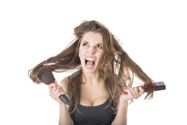 Mujer joven gritando mientras peina el pelo castaño por cepillo. Aislado sobre fondo blanco
 - Foto, Imagen