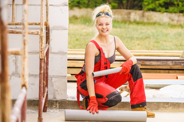 Mujer en dungarees llevando e instalando tubos de plástico en el sitio de construcción de su casa, construyendo una nueva casa
. - Foto, Imagen