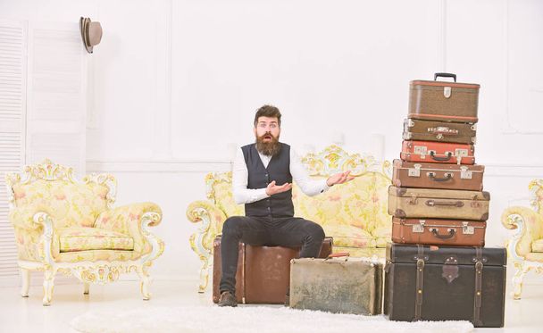 Macho elegant on surprised face sits, confused at end of packing, near pile of vintage suitcases. Luggage and relocation concept. Man with beard and mustache packed luggage, white interior background - Foto, afbeelding