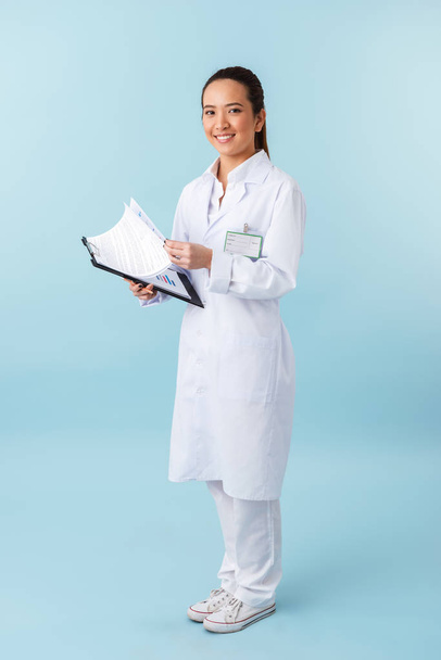 Photo of a cheerful young woman doctor posing isolated over blue wall background holding clipboard. - Foto, afbeelding