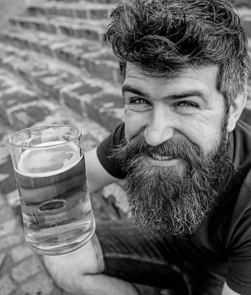 Guy having rest with draught beer. Hipster on cheerful smiling face drinks beer outdoor. Celebration concept. Man with beard and mustache holds glass with beer while sits on stone stairs, defocused - Foto, Bild