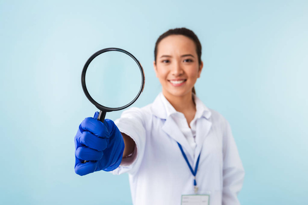Photo of a young woman doctor posing isolated over blue wall background holding magnifying glass. - Фото, изображение