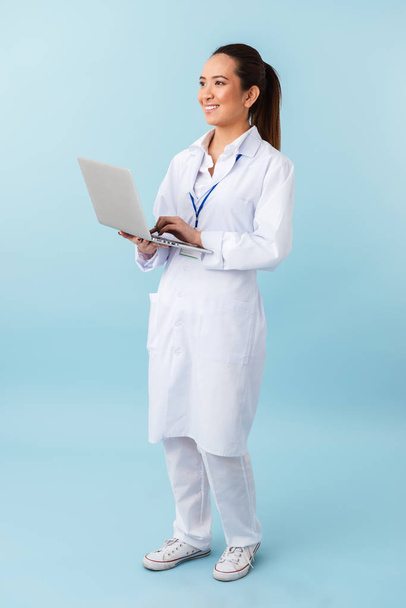 Photo of a young woman doctor posing isolated over blue wall background using laptop computer. - 写真・画像