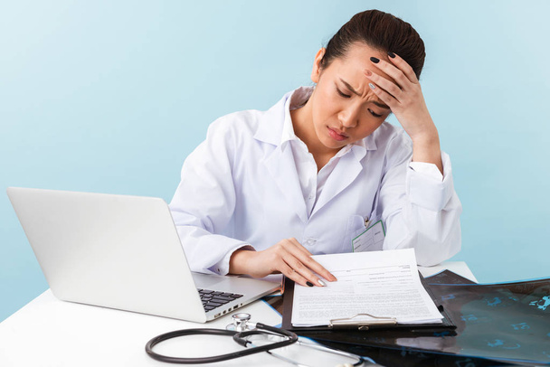 Photo of a young tired bored woman doctor with headache posing isolated over blue wall background using laptop computer. - Foto, immagini