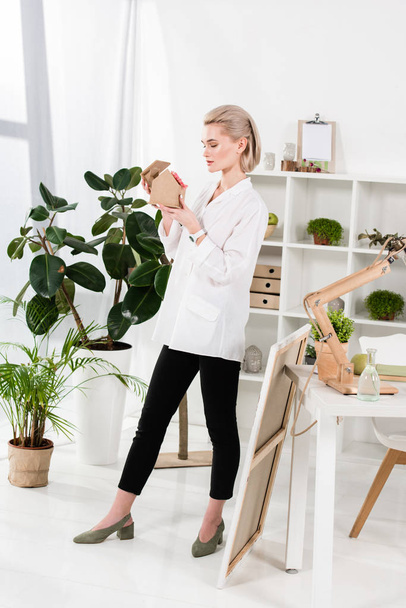 businesswoman holding small cardboard house with flower near green plants in office, environmental saving concept  - Photo, Image