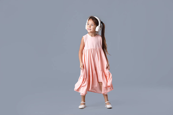 cute smiling little child girl in pink dress and headphones listens to music and dances on gray background. - 写真・画像