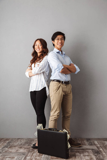 Full length of excited asian couple standing over gray background, holding briefcase full of money banknotes - Foto, imagen