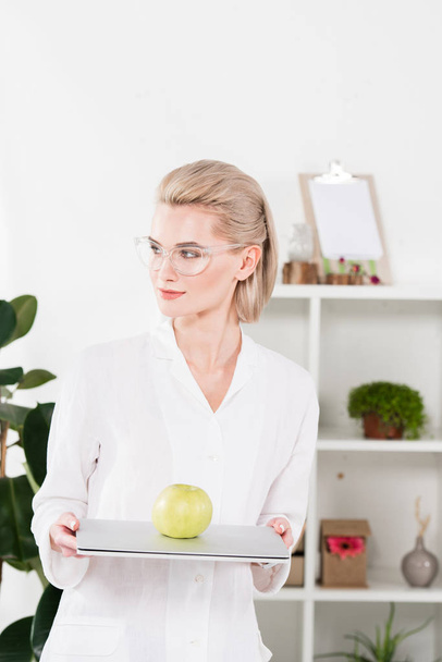 atractiva mujer en gafas de pie con portátil y manzana verde en la oficina
  - Foto, Imagen