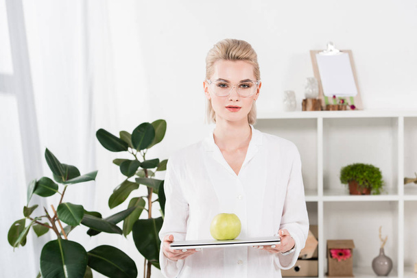 attractive businesswoman in glasses holding laptop with green apple in office  - Foto, afbeelding
