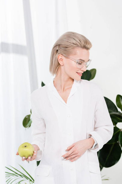 cheerful businesswoman in glasses holding green apple in office  - Foto, Imagen