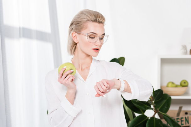 bella donna d'affari in occhiali guardando orologio mentre tiene mela verde in ufficio
  - Foto, immagini